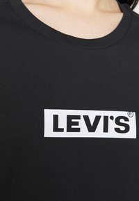 Černé bavlněné tričko s bílým obdélníkovým patche s logem Levi's v tučném černém písmu. Kulatý výstřih, hladká textura.