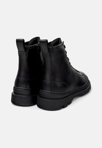 Une paire de bottes noires en cuir jusqu'à la cheville avec un laçage à l'avant et une fermeture éclair sur le côté, une semelle épaisse et robuste, et des languettes à l'arrière, présentées sous un angle arrière.