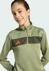 Mäta zelený športový top s vysokým golierom, oranžovými akcentmi a textúrovaným tmavozeleným horizontálnym pruhom cez hrudník.