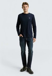 Marineblauw shirt met lange mouwen, ronde hals en klein logo, gecombineerd met donkerblauwe jeans en zwarte laarzen. Soepele stof, casual pasvorm.