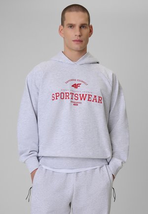 Młody mężczyzna w jasnoszarej bluzie z kapturem i dopasowanych spodniach dresowych z czerwonym napisem „SPORTSWEAR” na piersi, ręce w kieszeniach.