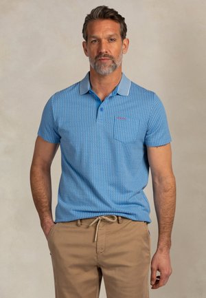 Man met zout-en-peper haar en baard, gekleed in een blauw poloshirt met patroon en beige broek met trekkoord, staand tegen een neutrale achtergrond.