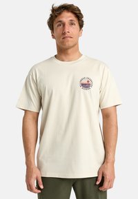 Camiseta de algodón de color beige con manga corta y escote redondo, que presenta un logotipo gráfico circular en el lado izquierdo del pecho.