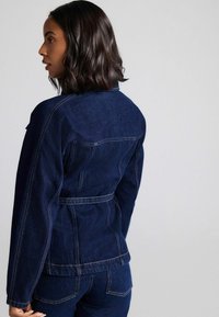 Kvinde med skulderlangt mørkt hår iført en mørkeblå denimjakke og matchende jeans, der vender til venstre mod en ensfarvet baggrund.