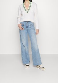Weißer Kabelstrickpullover mit V-Ausschnitt und neongrünen Streifen, kombiniert mit hoch taillierten, hellblauen weiten Jeans und gemusterten Sneakers.
