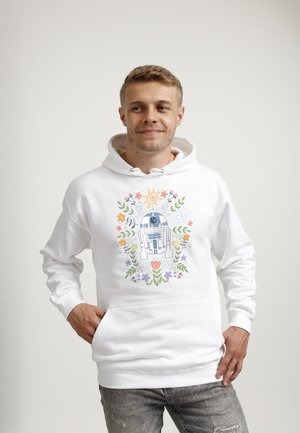 Joven con sudadera blanca con capucha con diseño colorido de robot R2-D2 y flores, de pie con la mano en el bolsillo, sonriendo ligeramente, con fondo liso.