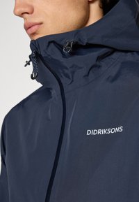 Gros plan sur une personne portant une veste Didriksons bleu marine avec une fermeture éclair et des cordons de serrage ajustables pour la capuche.