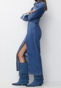 Jeansklänning med långa ärmar, sprund framtill och strukturerad silhuett. Kombinerad med slappa, midikorta blå jeansstövlar.