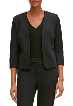 Frau in einem schwarzen taillierten Blazer mit Dreiviertelärmeln über einem schwarzen V-Ausschnitt-Top und passenden schwarzen Hosen.