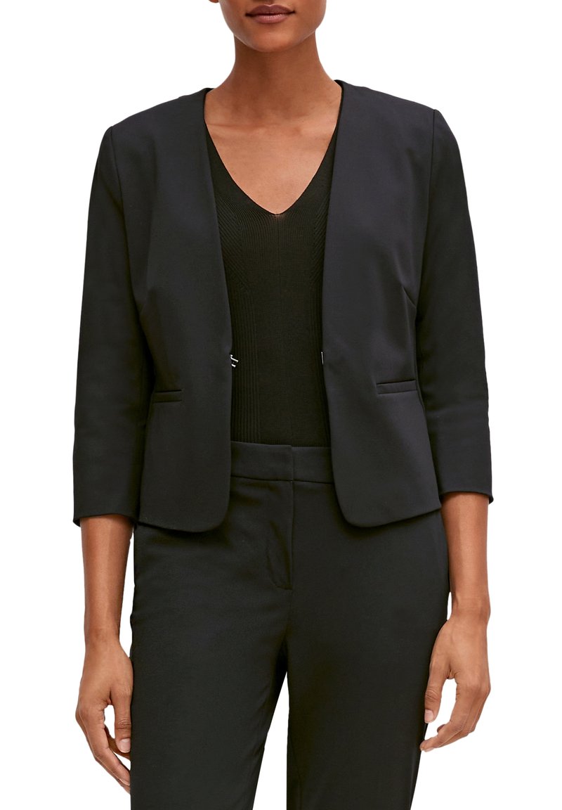 Vrouw in een zwart op maat gemaakt blazer met driekwart mouwen over een zwart V-hals top en bijpassende zwarte broek.