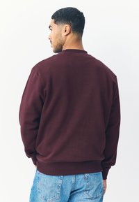 Vinröd sweatshirt med ribbade muddar och fåll, mjuk textur, rund halsringning, visas bakifrån mot en enfärgad vit bakgrund.