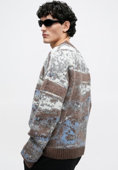 Pleteni pulover z teksturiranim vzorcem v odtenkih rjave, sive in modre barve. Klasičen kroj z rebrastim robom in manšetami. Model nosi sončna očala.