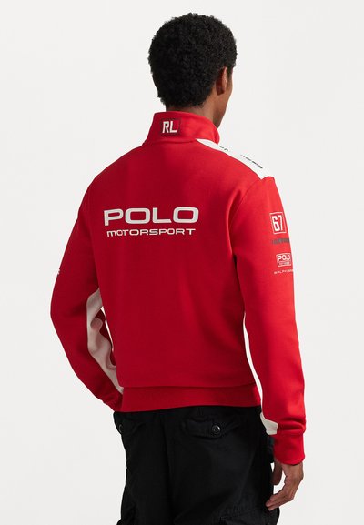 Červený sveter s vysokým golierom, s bielymi akcentmi na pleciach a s textom "POLO MOTORSPORT" výrazne zobrazeným na chrbte.