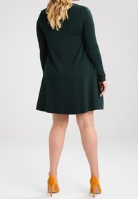 Zielona sukienka z długimi rękawami o rozszerzonym fasonie, gładkiej teksturze i długości do kolan. W połączeniu z jaskrawymi pomarańczowymi szpilkami.