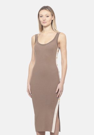 Giovane donna che indossa un vestito taupe aderente senza maniche con pannelli laterali bianchi e bottoni, in piedi contro uno sfondo chiaro e uniforme.