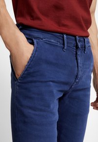 Blå bomullbyxor med slim fit, med sidofickor, bälteshällor och en mjuk yta. Bärs med en vinröd skjorta.