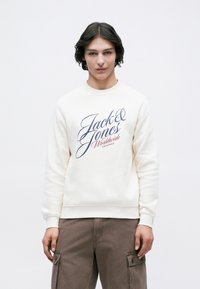 Mladý človek s tmavými vlasmi, oblečený v krémovej mikine s nápisom "Jack & Jones Worldwide Originals" a hnedých cargo nohaviciach, stojí na bielom pozadí.