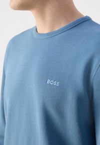 Kék bordázott pulóver, kerek nyakkivágással és kis fehér "BOSS" logóval a mellkason. Texturált anyagból készült.