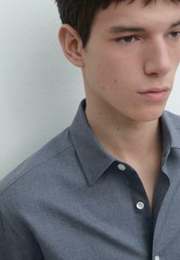 Šedá košile na knoflíky s měkkou texturou, s límečkem a jedním bílým knoflíkem u krku. Detail na modelčin hrudník a tvář.