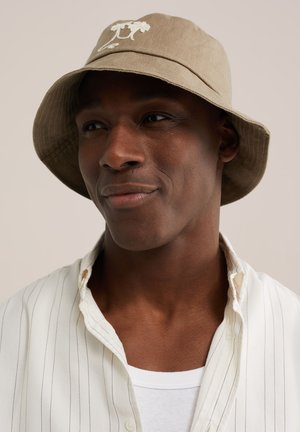 Uomo dalla pelle scura che indossa un cappello beige a tesa larga con ricami floreali, una camicia a righe bianche sopra una maglietta bianca, guarda di lato.