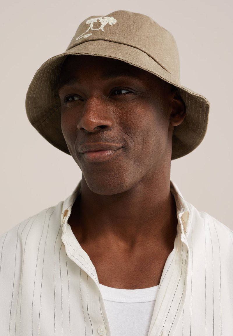 Homme à la peau foncée portant un chapeau bob beige avec une broderie florale, une chemise blanche à fines rayures portée sur un t-shirt blanc, regardant sur le côté.