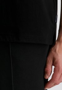 Nahaufnahme einer Hand, die neben einer schwarzen Hose und einem schwarzen Hemd mit einem kleinen "pierre cardin"-Label an der Seitennaht des Hemds ruht.