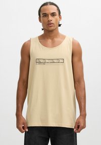 Joven con trenzas llevando una camiseta sin mangas beige con un mapa topográfico y el texto "White Peak" en el frente.