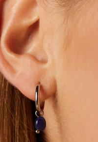 Boucle d'oreille en argent avec une perle bleue ronde suspendue, positionnée près d'une oreille humaine avec des cheveux fins visibles et une peau lisse.