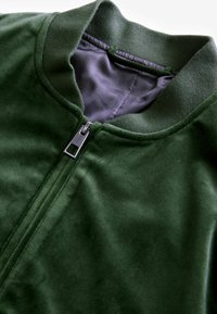 Giacca verde con zip caratterizzata da una texture morbida, colletto a coste e un interno a motivo herringbone contrastante. Dettagli con zip in metallo inclusi.