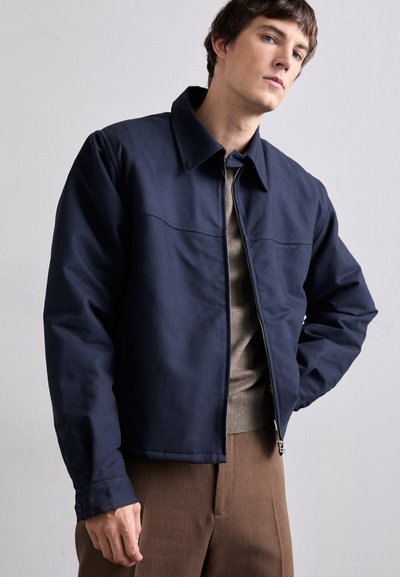 Navy blue jacket with a collared neck, zip closure, and front seam detail. Worn over a brown sweater and beige trousers.