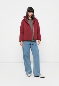 Burgunderfarbene Steppjacke mit Reißverschluss und Kapuze, getragen über einem grauen Oberteil, kombiniert mit lockeren blauen Jeans und schwarz-weißen Schuhen.