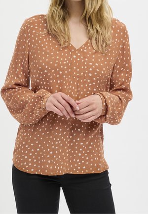 Kobieta w brązowej bluzce z długim rękawem w nieregularne białe groszki i czarnych spodniach, stojąca ze splecionymi palcami.