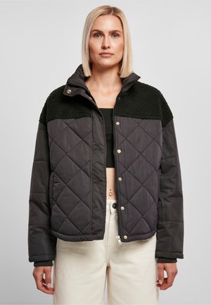 Kvinde med kort blondt hår iført en sort quiltet jakke med tekstureret øverste panel, sort crop top og hvide bukser mod en ensfarvet baggrund.
