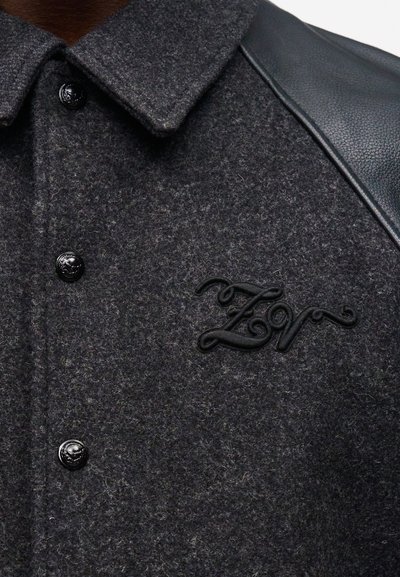 Σκούρο γκρι μάλλινο σακάκι με μαύρο δερμάτινο πάνελ στον ώμο, μαύρα κουμπιά και μαύρα κεντημένα αρχικά "ZN" στο στήθος.