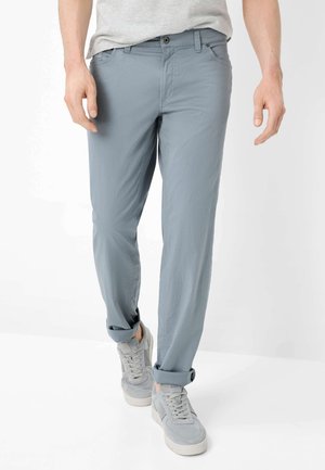 Uomo con pantaloni casual azzurri chiari risvoltati alle caviglie e scarpe da ginnastica grigie, in piedi davanti a uno sfondo bianco.