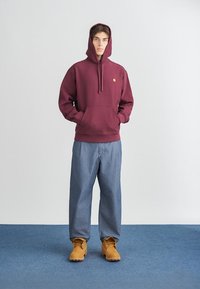 Bordeaux hoodie met een trekkoord en een zak aan de voorkant, gedragen met lichtblauwe denimspijkerbroek en tan laarzen op een blauwe tapijt achtergrond.