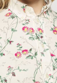 Blumen-Hemd mit Knopfverschluss, das einen cremefarbenen Hintergrund mit pinken und gelben Blumen sowie grünen Ranken zeigt. Es besteht aus strukturiertem Stoff und hat Knopfverschlüsse.