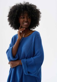 Blauwe gebreide trui met brede mouwen en een bootnek, met textuurpatronen en open steken langs de schoudernaad.