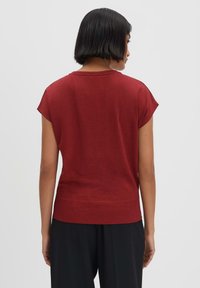 Top rosso a maniche corte realizzato in un tessuto morbido, con orlo a coste e vestibilità comoda, visto da dietro su uno sfondo chiaro.