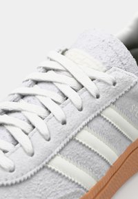 Vit mocka sneaker med texturerad ovandel, tre vertikala beige ränder och karameliserad gummisula. Har plana vita skosnören och logodetalj.