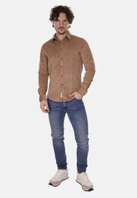 Camicia a maniche lunghe in corduroy marrone con bottoni, abbinata a jeans blu attillati e scarpe da ginnastica bianche con dettagli neri.