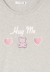 Tessuto grigio chiaro con testo bianco "Abbracciami," fiancheggiato da due cuori di glitter rosa e un orsetto di strass rosa tra di essi.