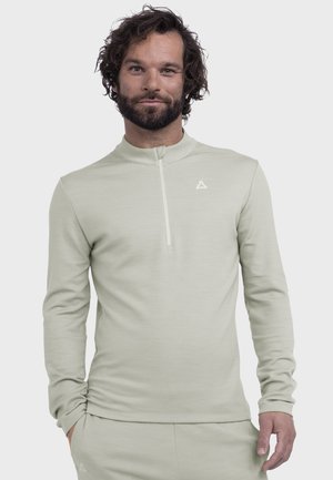 Mann mit Bart und lockigem Haar, der einen hellgrauen Langarmpullover mit halbem Reißverschluss und passende Hose vor einfarbigem Hintergrund trägt.
