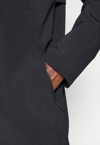 Μαύρο μπουφάν με κοντινή λήψη ενός χεριού σε τσέπη με φερμουάρ. Το ύφασμα φαίνεται λείο και ματ με μια μοντέρνα σχεδίαση.