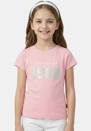 Fille souriante aux longs cheveux bruns portant un bandeau blanc, un t-shirt rose Lee Cooper avec le texte "1908", et un pantalon blanc sur un fond uni.