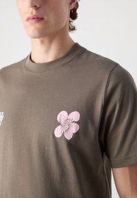 Olive green cotton t-shirt featuring a pink floral graphic on the left chest and a crew neckline. Short sleeves with a smooth texture.
