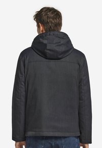 Man med en mörkgrå huva jacka, stående med ryggen vänd mot kameran, blå jeans synliga vid höfterna.