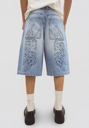 Személy világoskék farmer rövidnadrágot visel fekete törzsi lángmintákkal a hátsó zsebeknél és a lábakon, fehér zoknit és fekete cipőt.