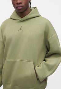 Jeune personne portant un sweat à capuche vert avec un petit logo Jumpman sur la poitrine, mains dans la poche avant, fond neutre.