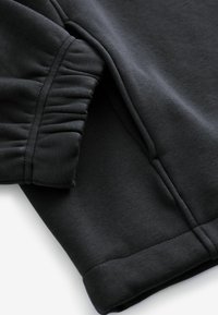 Čierny sweatshir tepela s nariasenými detailmi na manžete. Hladká textúra, elastický pás a kontrastné šitie pozdĺž švov.